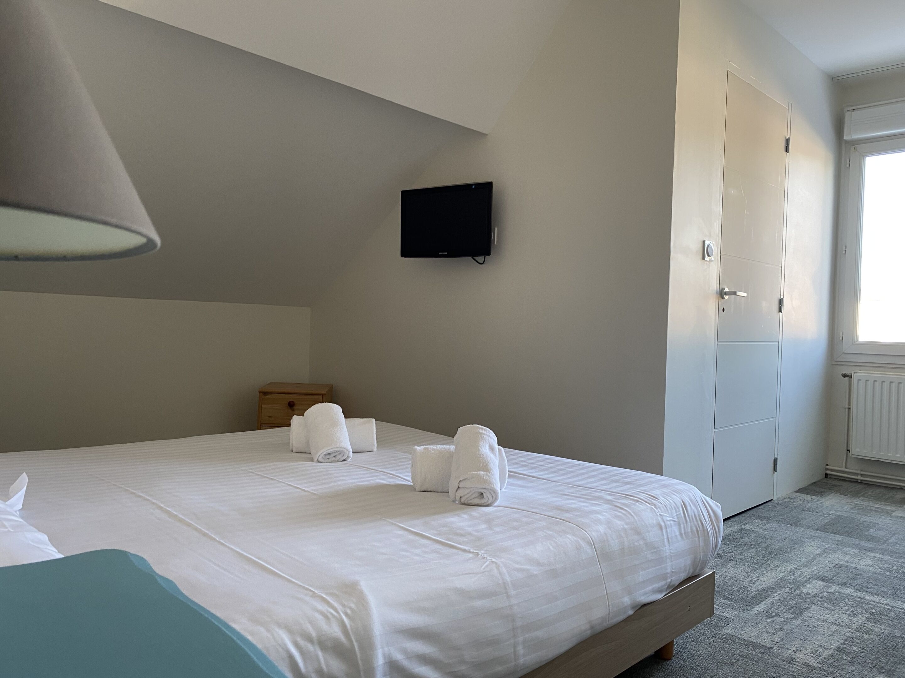 A hotel room with a bed, a flat-screen TV mounted on the wall, a wooden nightstand, and a door.