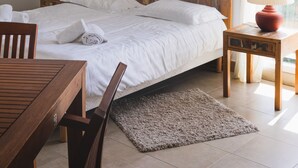 Une chambre à coucher avec une tête de lit en bois, un lit recouvert de draps blancs, une table en bois, une chaise, une petite table de chevet avec une lampe et une fenêtre avec des rideaux.