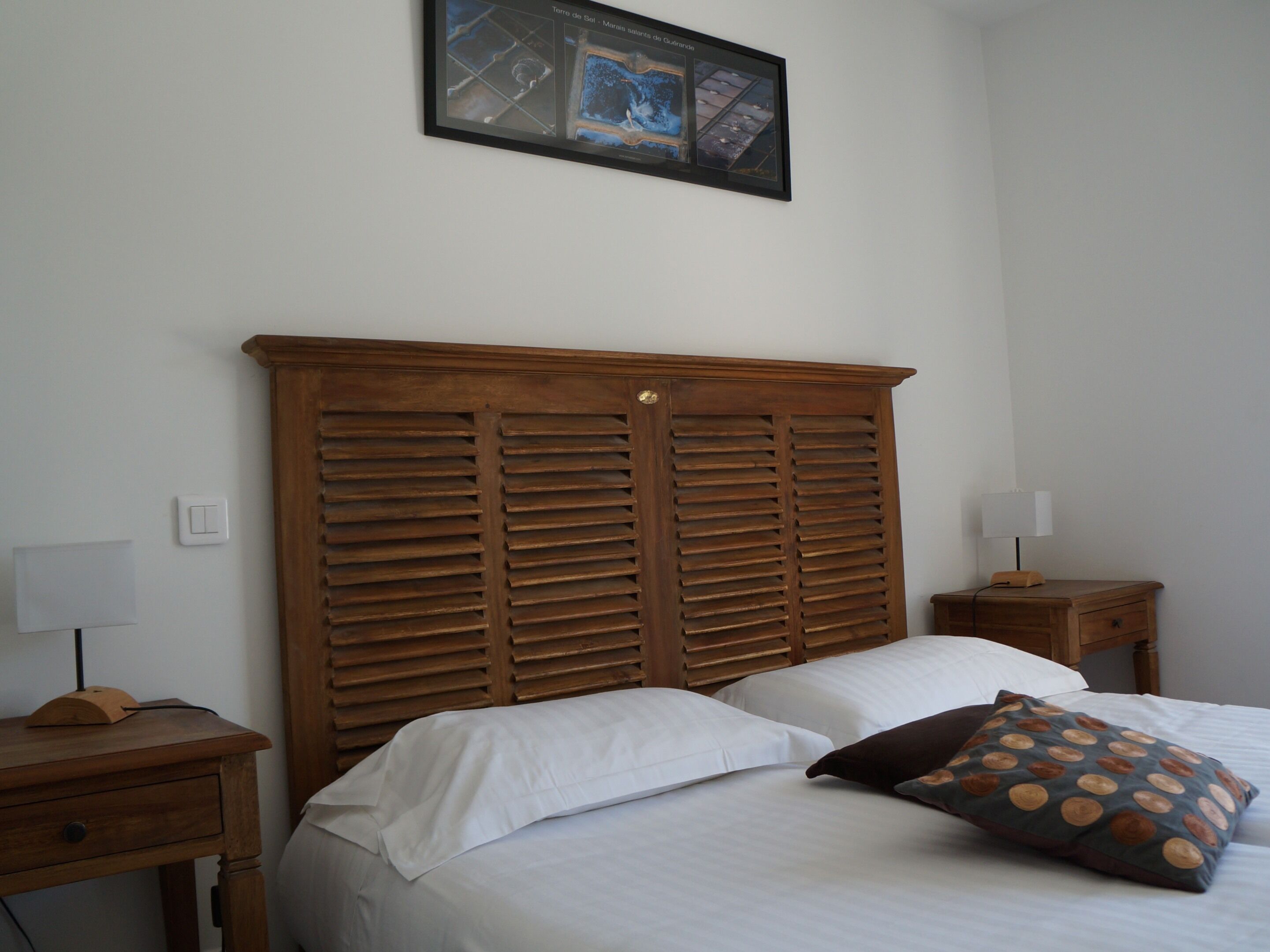 Un dormitorio con cabecera de madera, cama con sábanas blancas, mesita de noche con lámpara y un cuadro colgado en la pared.