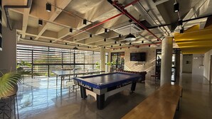 Une table de billard, une table de jeu de flipper et un banc en bois dans un espace intérieur moderne, avec des plafonds en béton et de grandes fenêtres.
