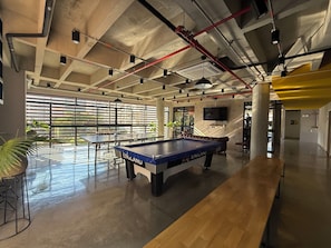 A billiards table, a pool table, and a wooden bench in a modern indoor space with concrete ceilings and large windows. - Flora Traveller House (Cali)