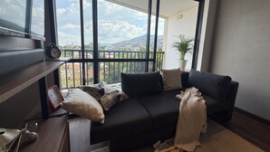 Un salon moderne avec un canapé noir, une petite table, une bibliothèque et une grande fenêtre donnant sur la ville.
