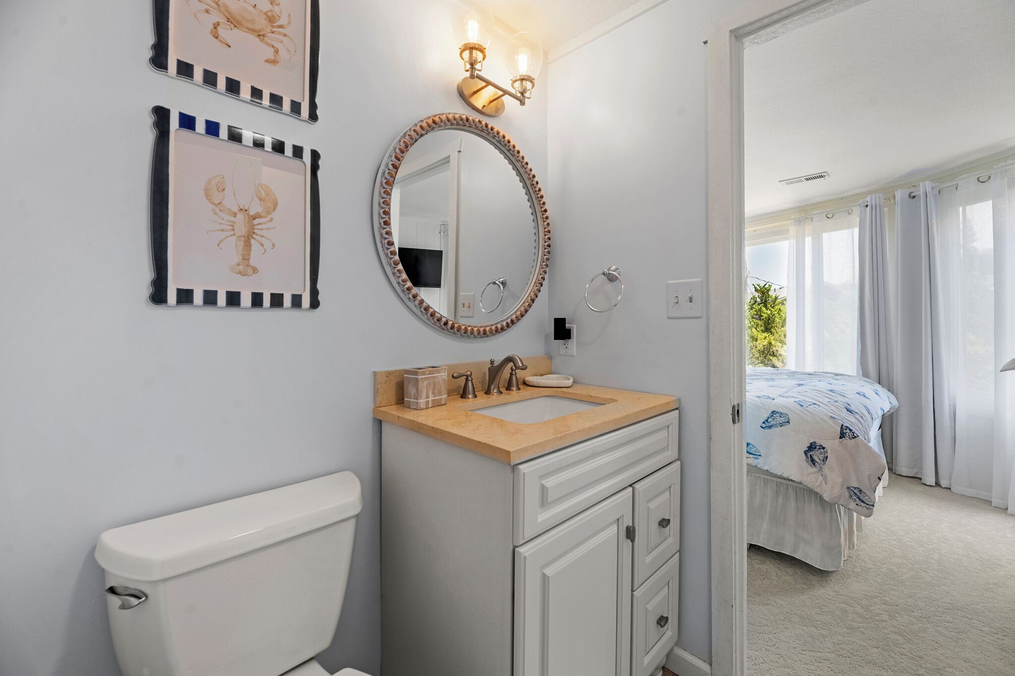 Une salle de bain avec des toilettes, un lavabo et un miroir. La pièce adjacente contient un lit recouvert d’une couverture bleue et blanche.