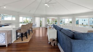 A spacious living area with a ceiling fan, a blue sofa, a kitchen area with a white island, and large windows offering a view of the outside. - Short Walk to Beach | Outdoor Entertainment Area (Salvo)