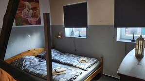 Une chambre avec deux lits, une fenêtre avec un store, une petite étagère et une lampe sur une table.
