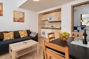 Un espace de vie compact comprenant un canapé, une table à manger avec des chaises, un coin cuisine et une petite table à manger agrémentée d’un saladier de fruits et de verres à vin.