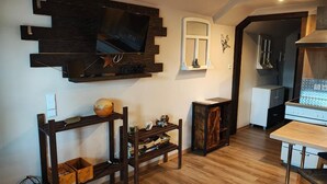 Ein Wohnzimmer mit an der Wand montiertem Fernseher, einem Esstisch und einem durch eine offene Tür sichtbaren Küchenbereich.