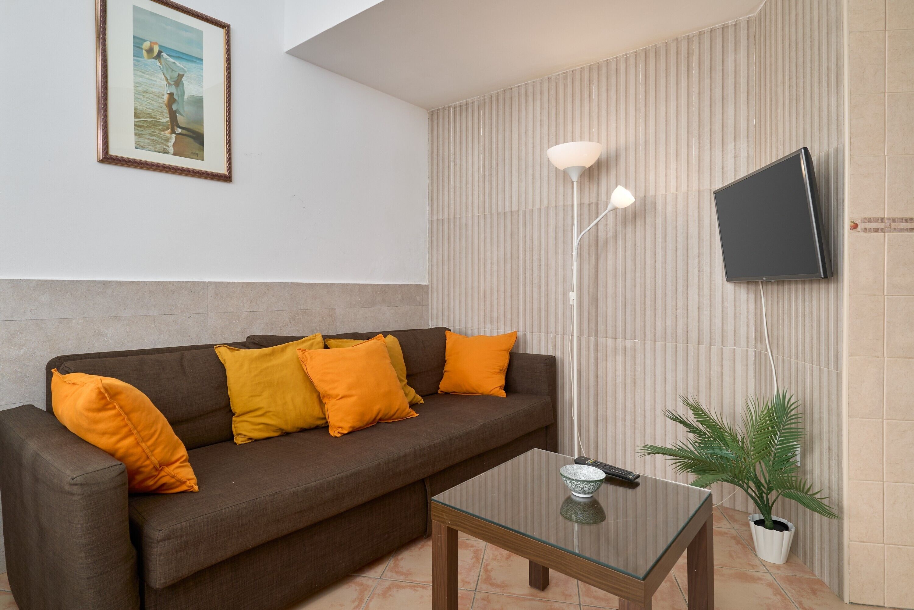 A living room with a brown sofa, orange cushions, a glass coffee table, a mounted TV, a floor lamp, and a potted plant.