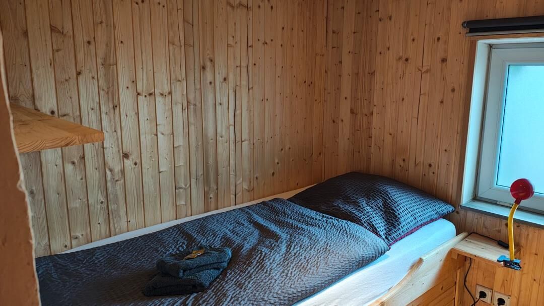 A small wooden room with a bunk bed, a window with a view, and a small bedside table with a lamp.