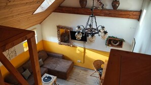 A room with a sloped ceiling, a wooden ceiling beam, a brown sofa, a wicker chair, a table with a plate, and a chandelier. - Modern loft apartment No. 3 with terrace (Aurich)