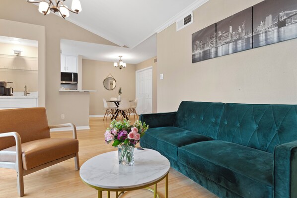 A living room with a teal sofa, a brown armchair, a round coffee table with a vase of flowers, and a cityscape painting on the wall.