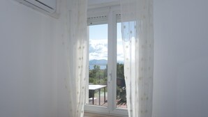 Ein Zimmer mit Fenster, weißen Vorhängen und Blick auf eine Terrasse mit Tisch und Stühlen.