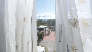 Blick durch ein Fenster mit transparenten Vorhängen, der eine gepflasterte Außenfläche mit überdachter Sitzgruppe und Grünpflanzen zeigt.