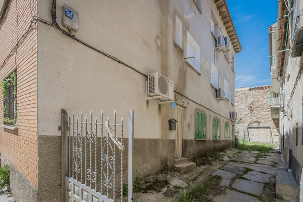 A narrow alleyway with a building featuring a metal gate, air conditioning units, and a stone wall.