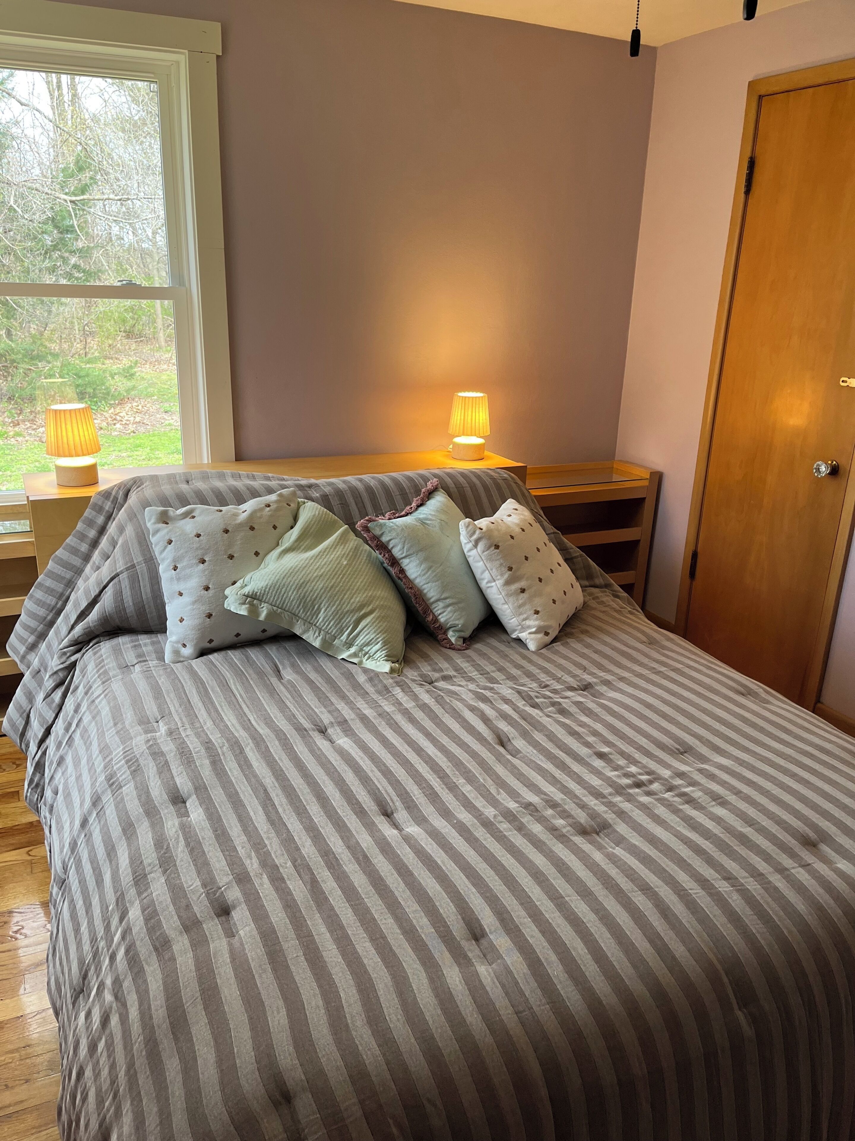 Una cama con ropa de cama a rayas, dos lámparas de noche, una mesita de noche de madera y vistas a árboles a través de una ventana.