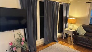 Una sala de estar con televisor de pantalla plana, cortinas oscuras, una mesita auxiliar blanca con una lámpara, un sofá de cuero marrón y una alfombra gris.