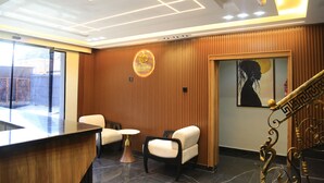 A reception area with a wooden panel wall, a reception desk, two chairs, a small round table, and a painting on the wall. - Victoria Plus Hotel & Suites (Asaba)
