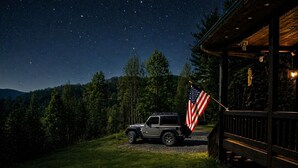 Ein Fahrzeug vor einer Holzhütte, eine amerikanische Flagge weht, umgeben von bewaldeter Landschaft unter einem sternenübersäten Nachthimmel.