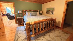 Ein Bett mit Holzrahmen und einem gemusterten Bettbezug, ein Holz-Kopfstück, ein kleiner Nachttisch mit Lampe und ein großes Fenster mit Jalousien.