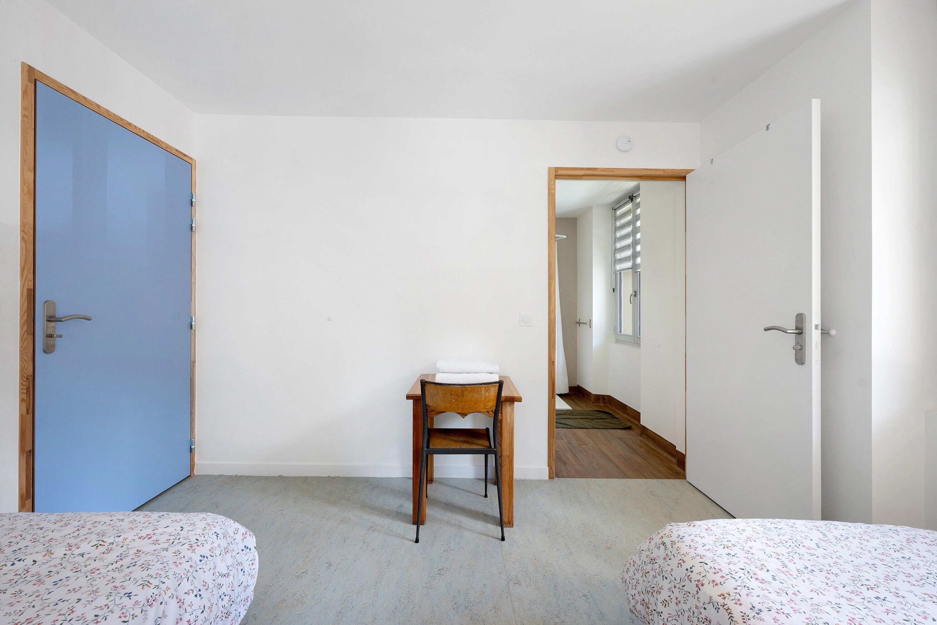 Una habitación con una puerta azul, una silla de madera, una cama con ropa de cama estampada y un pasillo que conduce a otra habitación.