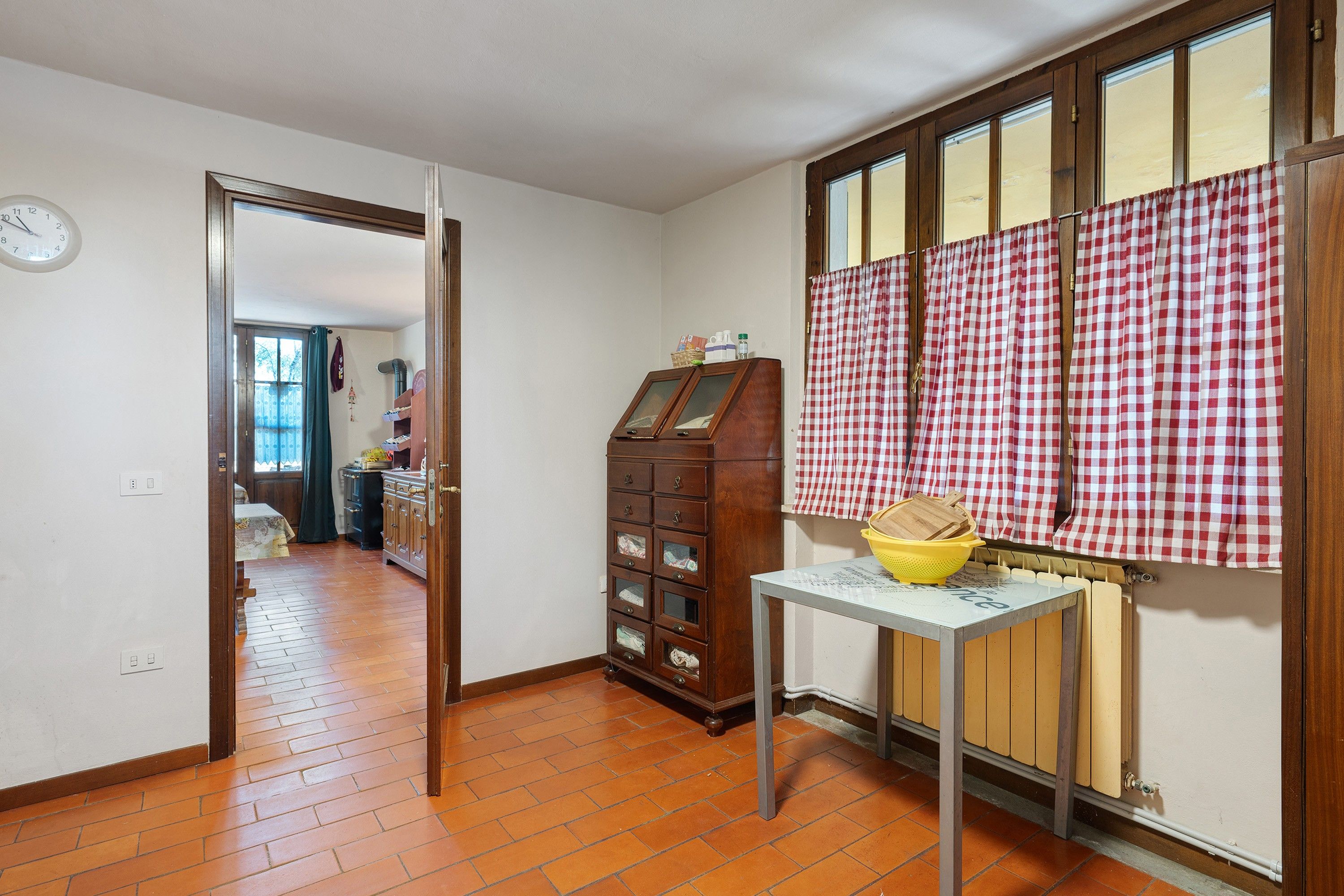 A kitchen with a wooden cabinet, a table with a yellow bowl, and a window with checkered curtains.