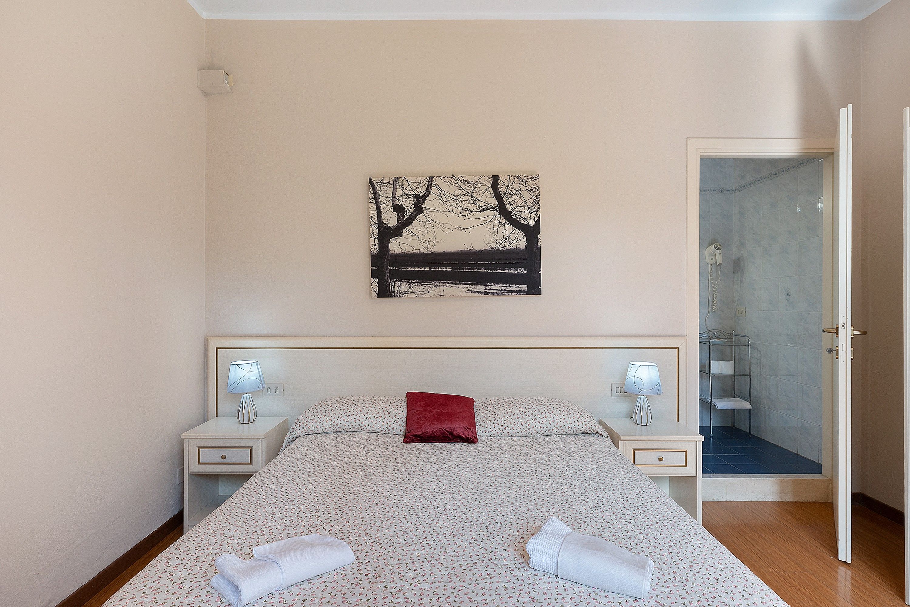 A bedroom with a bed, two bedside tables, a nightstand, a red pillow, a wall-mounted picture, and a bathroom door.