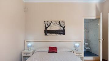 A bedroom with a bed, two bedside tables, a nightstand, a red pillow, a wall-mounted picture, and a bathroom door.