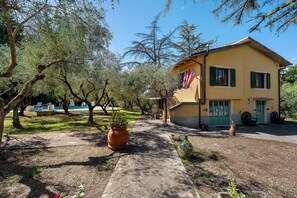 A yellow building with a green door and windows, surrounded by trees and a paved path. - Room 'Acqua' with Shared Pool, Communal Garden and Wi-Fi (Le Vedute)