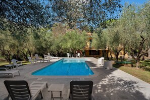 Una piscina con sillas de descanso y mesas, rodeada de árboles y un edificio al fondo.