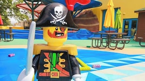 LEGO-merirosvo, jolla on pääkallokoristeinen hattu ja jonka kädessä on miekka, seisoo leikkikentällä, jossa on pöytiä ja aurinkovarjoja.
