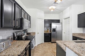 A kitchen with dark cabinets, stainless steel appliances, granite countertops, and white walls. - 3/2 Cozy Home Near UNT & Twu-pool, Bbq, Gym, Games (Denton)