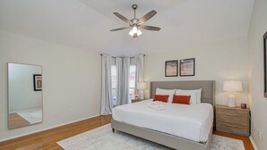 A bedroom with a bed, a ceiling fan, a nightstand, a lamp, a mirror, and a window with curtains. - 4BR Game Room + Firepit Sleeps 9 Near Eats (Houston)