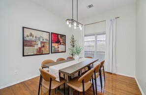 A dining area with a long table, six chairs, a vase with flowers, and two framed artworks on the wall. - 4BR Game Room + Firepit Sleeps 9 Near Eats (Houston)