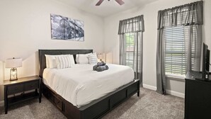 A bedroom with a bed, nightstand, ceiling fan, artwork, windows with curtains, and a TV stand. - Heart of Ferris: 3-bd/2-ba With Pool, Patio & BBQ (Ferris)