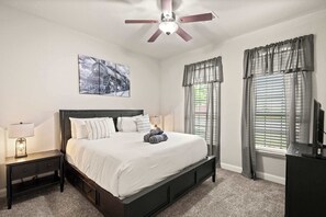 A bedroom with a bed, nightstand, ceiling fan, artwork, windows with curtains, and a TV stand. - Heart of Ferris: 3-bd/2-ba With Pool, Patio & BBQ (Ferris)