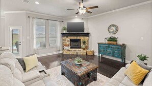 A living room with a stone fireplace, a ceiling fan, a television, and a blue cabinet. - Heart of Ferris: 3-bd/2-ba With Pool, Patio & BBQ (Ferris)