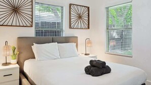 A neatly arranged bedroom with a large bed, two bedside tables with lamps, and decorative wall art above windows. - Serene Escape - Peaceful 3-bd Retreat With Yard (Grand Prairie)