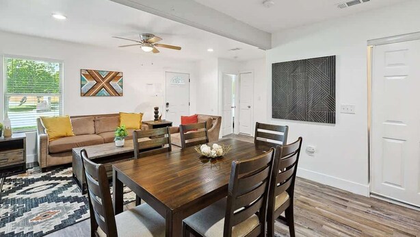 A spacious living room with a wooden dining table, comfortable seating, and a large window with blinds. - Serene Escape - Peaceful 3-bd Retreat With Yard (Grand Prairie)