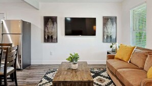 A modern living room with a wooden coffee table, a sofa, and a dining area with chairs. There are three framed artworks on the wall and a window with blinds. - Serene Escape - Peaceful 3-bd Retreat With Yard (Grand Prairie)