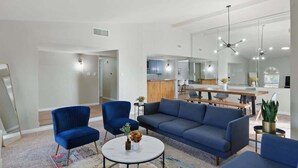 Uma sala de estar moderna com um sofá, cadeiras e uma mesa de centro. É visível uma cozinha ao fundo, com uma mesa de jantar.