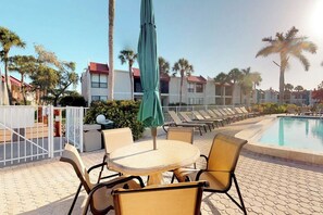 A poolside area with lounge chairs and a table, surrounded by palm trees and buildings. - Dans Florida Condos Vista Bay at Runaway Bay (Bradenton Beach)