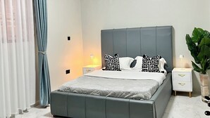 Un dormitorio con cama, dos mesitas de noche, una mesita auxiliar, una planta en maceta y una ventana con cortinas.