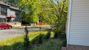 Una strada residenziale con un'auto rossa parcheggiata sul lato, un vialetto che conduce a una casa e un'area giardino con diverse piante.