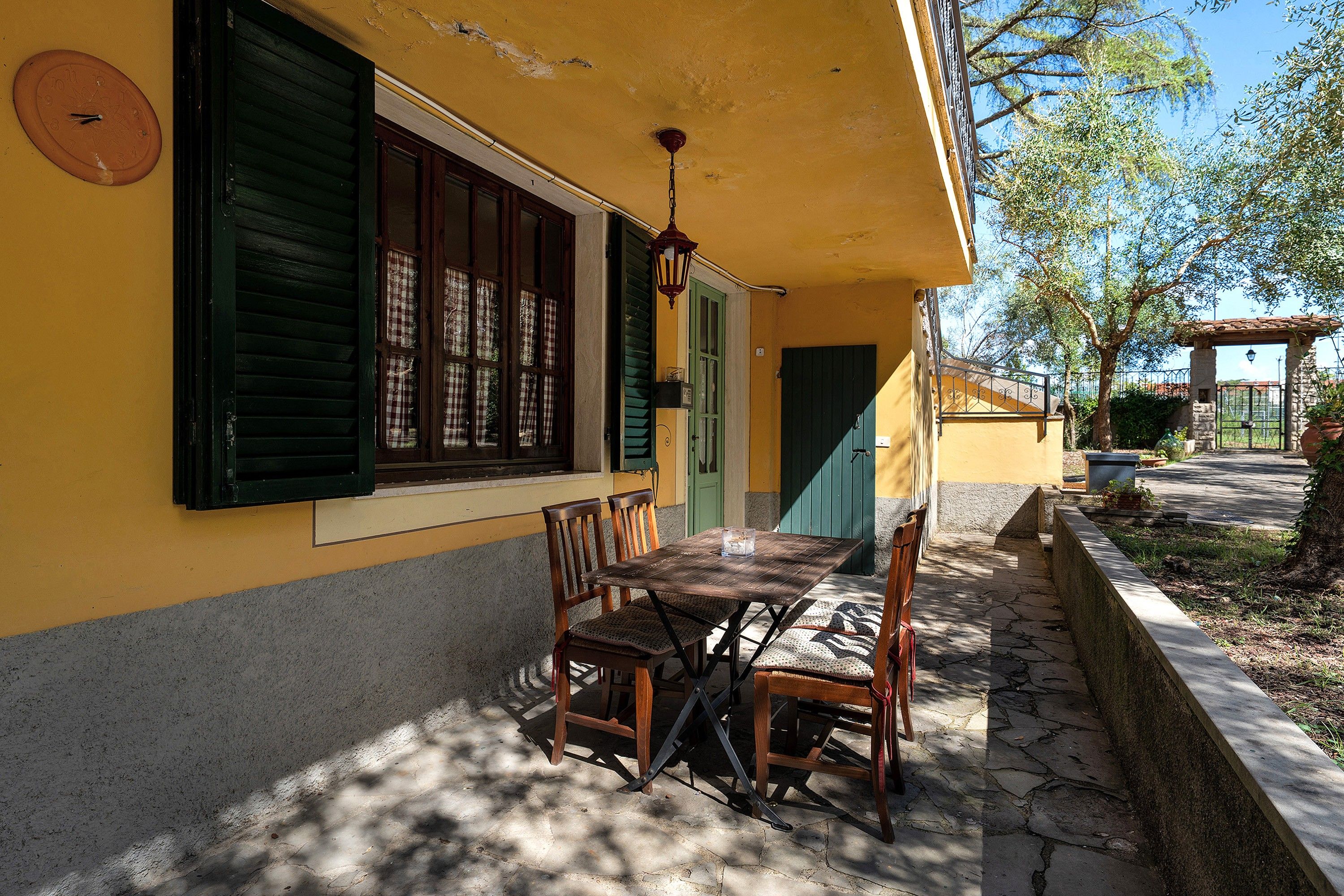 A patio with a table and chairs, a clock on the wall, and a door with green shutters.