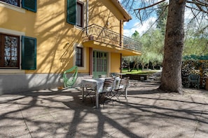 Un edificio amarillo con balcón, mesas al aire libre y un árbol.