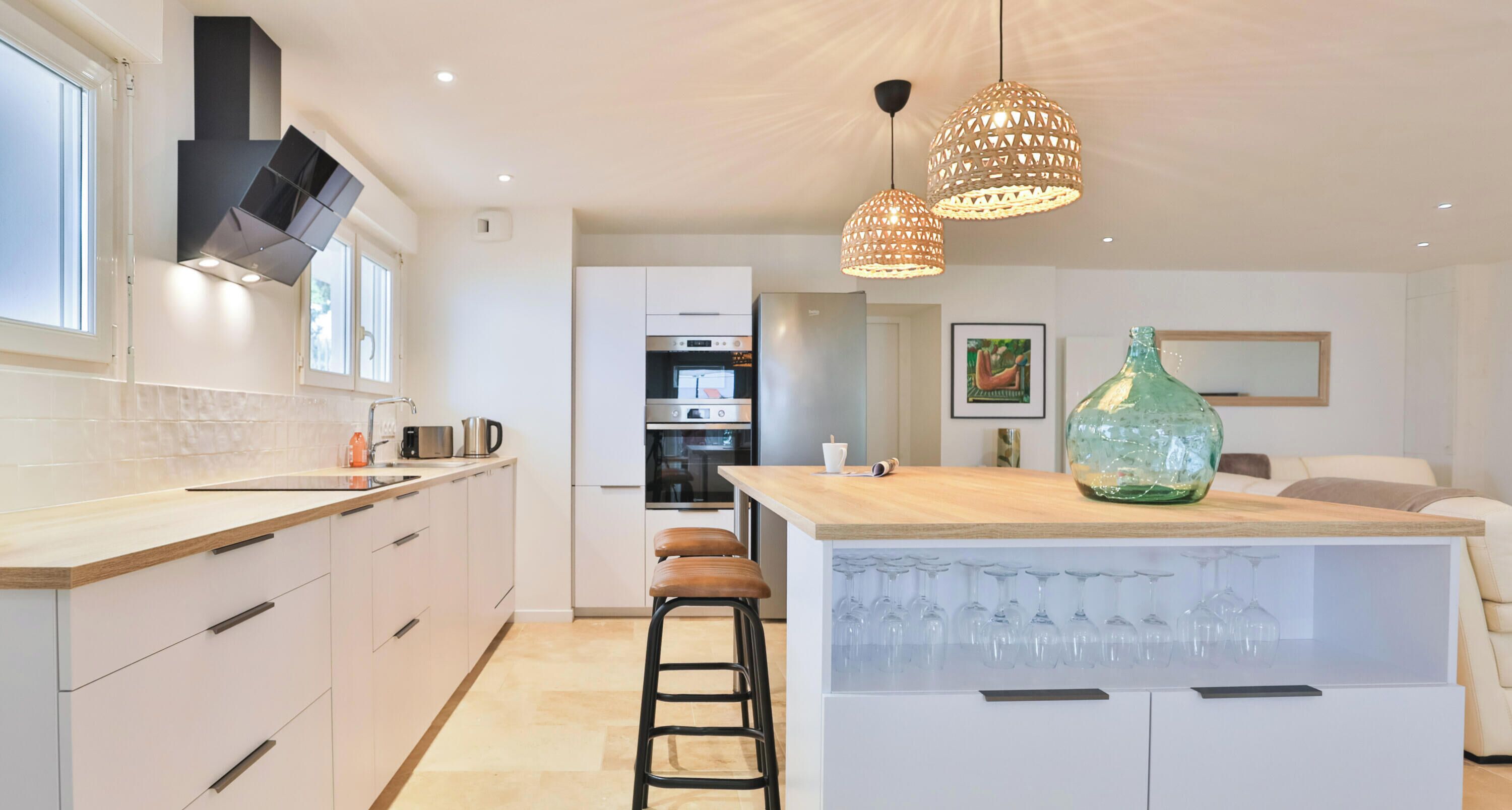 A modern kitchen with white cabinets, a wooden countertop, and a central island with stools. There are pendant lights above the island, and a large vase on top.