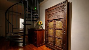 Un escalier en colimaçon, une porte en bois ornée de sculptures complexes et un meuble en bois sur lequel est posé un vase rempli de tournesols.