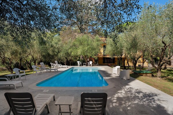 A swimming pool with lounge chairs and tables, surrounded by trees and a building in the background.