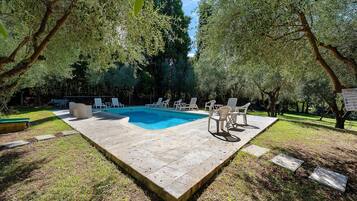 Piscina retangular com esguicho, deck ladrilhado, cercada por grama e árvores, com cadeiras de descanso e uma mesa.