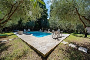 A rectangular swimming pool with a tiled deck, surrounded by grass and trees, with lounge chairs and a table.